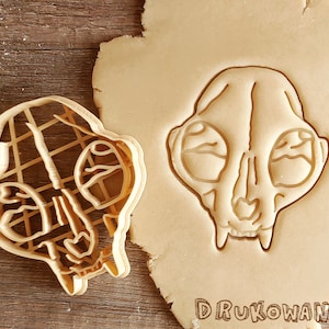 May include: A close-up of a cookie cutter in the shape of a cat skull. The cutter is made of plastic and has a detailed design. The cutter is on top of a piece of cookie dough.