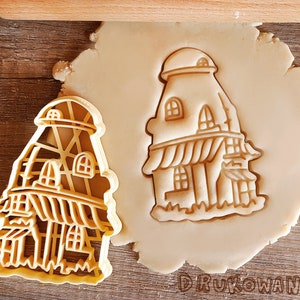 Peut inclure: Un emporte-pièce en forme de maison en pain d'épices avec un design détaillé. L'emporte-pièce est en plastique jaune et est présenté sur une feuille de pâte à biscuits.