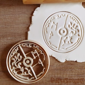 Peut inclure: Un emporte-pièce en bois en forme de bouclier rond avec un motif stylisé d'un arbre et les lettres "MA" dessus. L'emporte-pièce est sur une surface blanche.