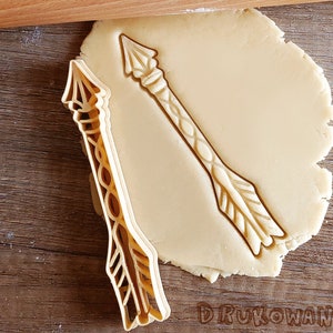 Könnte beinhalten: Ein goldfarbener Ausstecher in Form eines Pfeils mit filigranen Details. Der Ausstecher liegt auf einem Stück ungebackenem Keksteig.