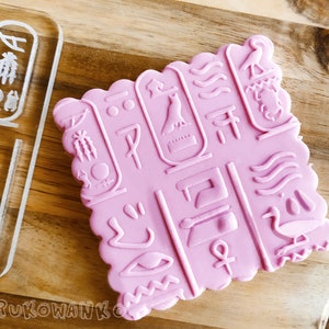 Puede incluir: Sellos de silicona rosa con un diseño de jeroglíficos egipcios. El sello presenta varios símbolos, incluyendo un pájaro, un ojo y un sol.