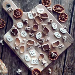 Può includere: Un set di 30 tagliabiscotti in plastica bianca a forma di varie rune e simboli su una superficie di legno.