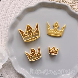 Può includere: Quattro tagliabiscotti in plastica color oro a forma di corone. I tagliabiscotti sono disposti su un piatto grigio con un bordo ondulato.