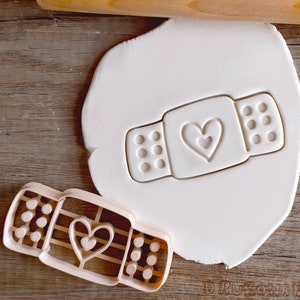 May include: A plastic cookie cutter in the shape of a plaster with a heart in the centre. The cutter is on a wooden surface with a white fondant imprint of the cutter.