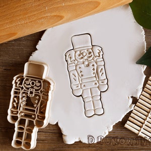 Könnte beinhalten: Ein beige Kunststoff-Nussknacker-Ausstecher und ein Abdruck in weißem Teig. Der Ausstecher hat die Form eines Soldaten mit Hut und Uniform. Im Hintergrund befindet sich ein Nudelholz aus Holz.