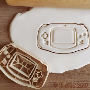 May include: A white plastic cookie cutter in the shape of a handheld video game console. The cutter is on a white surface.