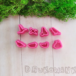 Pode incluir: Um conjunto de nove cortadores de biscoitos de plástico rosa em forma de pássaros. Os cortadores estão dispostos em duas filas sobre uma superfície de madeira branca.