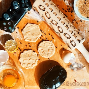 May include: A wooden rolling pin with a design of video game controllers and a game console. There are also several cookie cutters in the shape of video game controllers on a wooden surface.