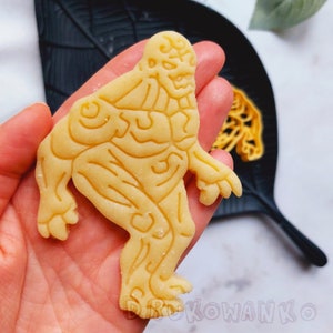 May include: A yellow cookie shaped like a monster with detailed muscle definition. The cookie is held in a hand.