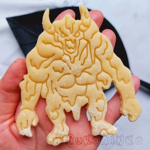 May include: A detailed cookie cutter in the shape of a monstrous creature with horns, a large mouth, and a muscular body. The cookie cutter is made of a light brown dough and is dusted with powdered sugar.