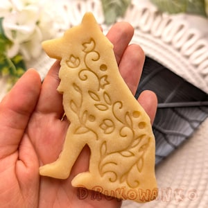 Puede incluir: Una galleta de color marrón claro con forma de perro con un diseño floral. La galleta se sostiene en una mano.