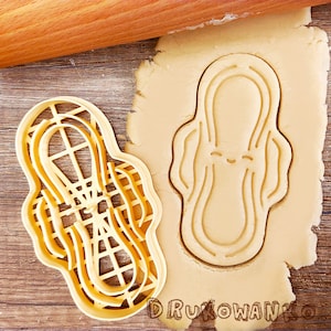 Puede incluir: Un cortador de galletas de plástico beige con forma de compresa, junto a un recorte en masa. Un rodillo de madera descansa en el fondo. La palabra "DRUKOWANKO" está impresa en la esquina inferior derecha.