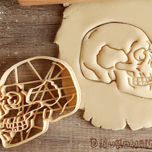 May include: A close-up of two cookie cutters shaped like a skull. The cookie cutters are made of a light brown plastic and have a detailed design. The cookie cutters are on a wooden surface.