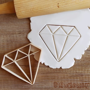 May include: A plastic diamond-shaped cookie cutter on a white surface with a rolling pin in the background. The cookie cutter is used to create diamond-shaped cookies.