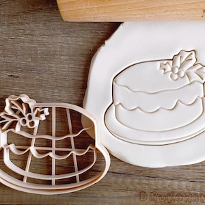 Könnte beinhalten: Ein weißer Plastik-Ausstecher in Form eines Weihnachtskuchens mit einer Stechpalme oben drauf. Der Ausstecher liegt auf einer weißen Oberfläche.