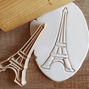 Puede incluir: Un cortador de galletas de plástico blanco con forma de la Torre Eiffel. El cortador está sobre una superficie blanca con un rodillo de madera en el fondo.