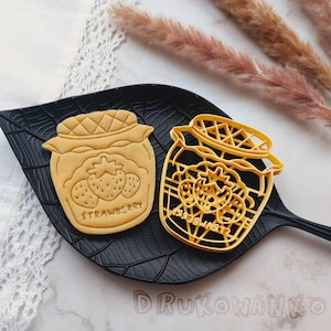 May include: Two yellow cookie cutters shaped like jars of strawberry jam. The jar on the left has the word "Strawberry" written on it. The jar on the right has a more detailed design with a grid pattern and a strawberry inside.