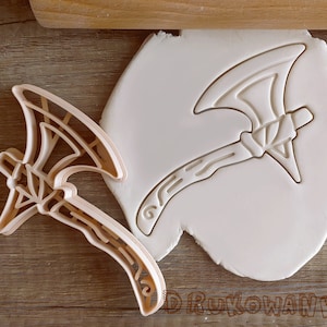 May include: A white plastic cookie cutter in the shape of an axe. The cutter is on a white surface.
