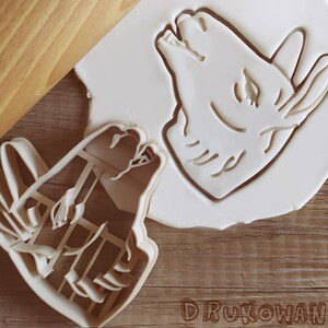 Pode incluir: Dois cortadores de biscoitos de pl&aacute;stico branco, um com a forma de cabe&ccedil;a de lobo e o outro com a forma de um lobo a uivar para a lua. Os cortadores est&atilde;o sobre uma superf&iacute;cie de madeira.