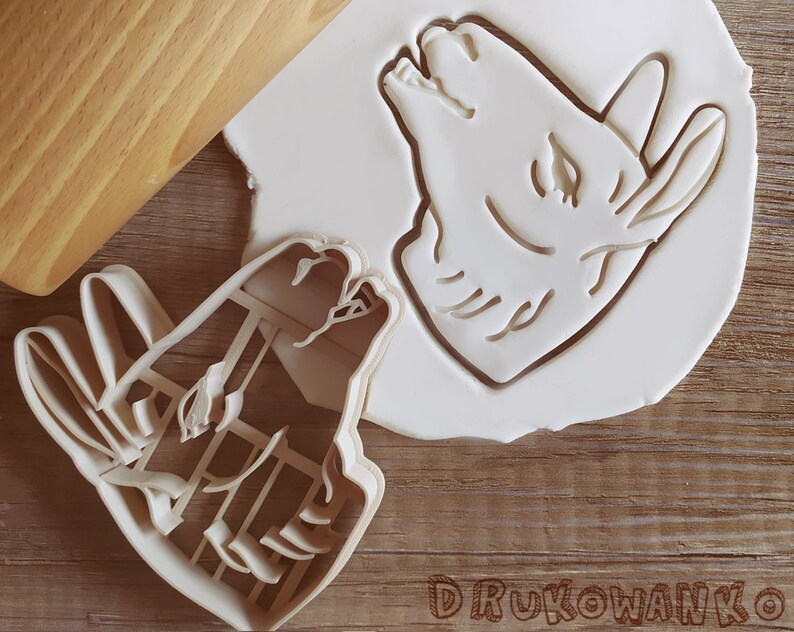 Pode incluir: Dois cortadores de biscoitos de pl&aacute;stico branco, um com a forma de cabe&ccedil;a de lobo e o outro com a forma de um lobo a uivar para a lua. Os cortadores est&atilde;o sobre uma superf&iacute;cie de madeira.
