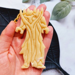 May include: A cookie cutter in the shape of a tree with a detailed design. The cutter is made of a light brown dough and is held in a hand.