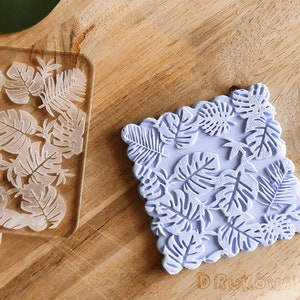 May include: Two silicone stamps with a tropical leaf design. The stamp on the left is clear and the stamp on the right is a light purple color.