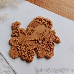 Puede incluir: Un sello de goma marrón con un diseño detallado de un pájaro posado en una rama con flores y bayas. El sello está diseñado para su uso en repostería y decoración.