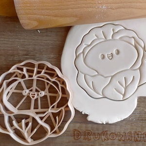 May include: A white plastic cookie cutter in the shape of a cabbage with a smiling face. The cutter is on a wooden surface with a rolling pin and a white background.