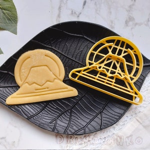 May include: Two yellow plastic cookie cutters on a black background. One cutter is shaped like Mount Fuji and the other is shaped like a Ferris wheel.