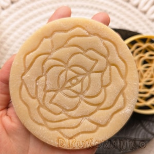 Puede incluir: Un cortador de galletas redondo de color beige con un diseño de rosa detallado. El cortador está hecho de un material liso y texturizado y tiene un borde elevado.