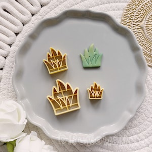 以下が含まれることがあります： 草の形をしたクッキーカッターが4つあります。3つは黄色で、1つは緑色です。カッターは灰色の皿の上にあります。