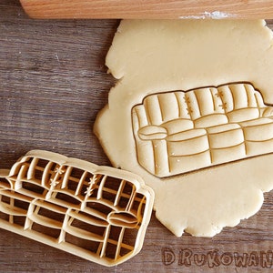 Op de afbeelding: Een koekjesvormpje in de vorm van een bruine bank met een lichtbruin kussen. Het koekjesvormpje ligt op een vel ongebakken koekdeeg.