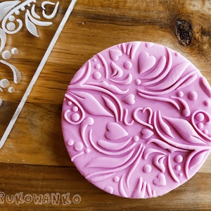 Puede incluir: Un sello de silicona rosa con un diseño en relieve de corazones, hojas y espirales. El sello está sobre una superficie de madera.