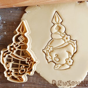May include: A cookie cutter in the shape of a cartoon character wearing a crown and a dress with flowers. The cutter is made of metal and has a shiny finish.
