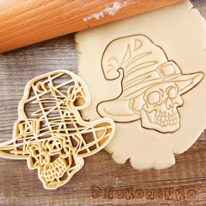 Puede incluir: Dos cortadores de galletas con forma de calavera con un sombrero de bruja. Los cortadores están hechos de plástico marrón claro y están sobre una superficie de madera.