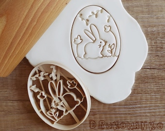Puede incluir: Cortador de galletas de plástico blanco en forma de huevo de Pascua con un conejito dentro. El conejito sostiene una flor. El cortador está sobre una superficie blanca.