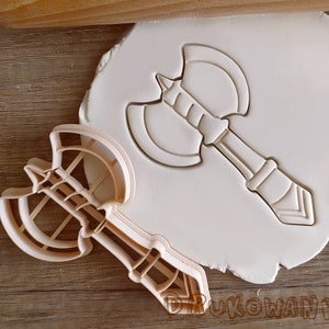 May include: A plastic cookie cutter in the shape of an axe. The cutter is on a white surface with a raised impression of the axe.