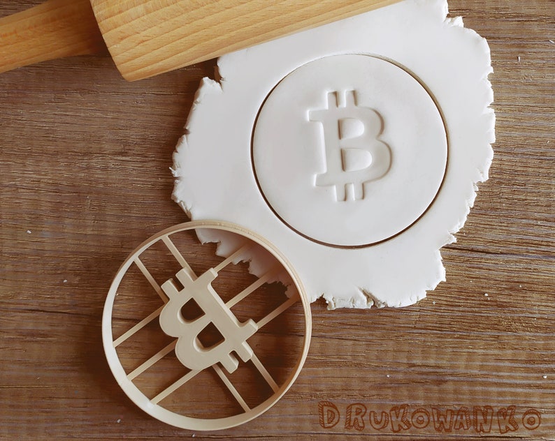 May include: A white cookie cutter in the shape of a Bitcoin symbol, with a rolling pin and a piece of white dough in the background.