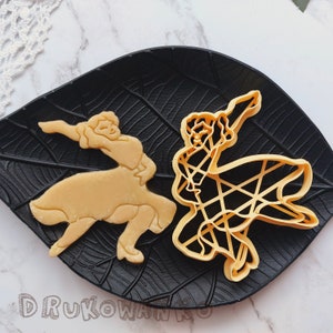 May include: Two cookie cutters in the shape of dancers. The cutter on the left is a solid shape of a woman in a dress. The cutter on the right is a more detailed outline of a woman in a dress. Both cutters are on a black leaf-shaped background.