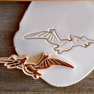 Peut inclure: Un emporte-pièce en plastique brun en forme de ptérodactyle, un dinosaure volant avec des ailes, un long cou et un bec. L'emporte-pièce est posé sur un rouleau à pâtisserie blanc, lui-même posé sur une surface en bois.
