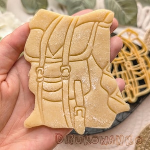 Puede incluir: Una galleta de color marrón claro con forma de mochila con un contorno detallado de correas y hebillas. La galleta está espolvoreada con azúcar en polvo.