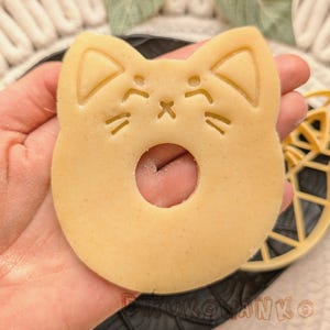 May include: A light brown cookie cutter in the shape of a cat's face with a hole in the middle. The cookie cutter has cat's ears, eyes, nose, and whiskers.