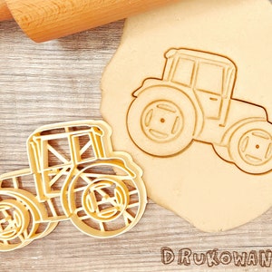 May include: A beige tractor cookie cutter next to a tractor-shaped cookie on a wooden surface. The cookie cutter is made of plastic and has a detailed design. The word "DRUKOWANKO" is printed at the bottom.