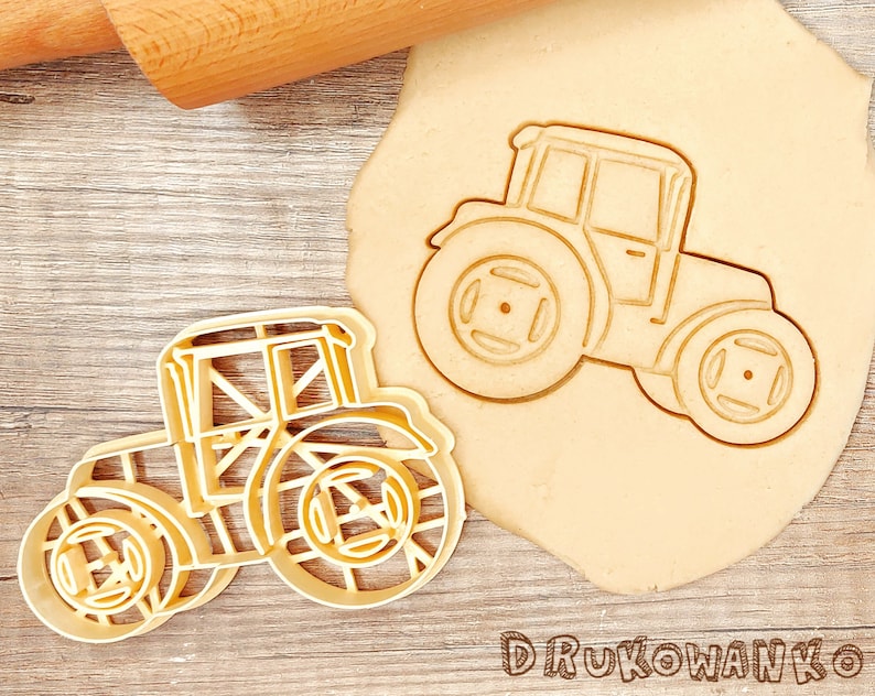 May include: A beige tractor cookie cutter next to a tractor-shaped cookie on a wooden surface. The cookie cutter is made of plastic and has a detailed design. The word "DRUKOWANKO" is printed at the bottom.