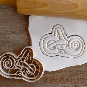 May include: A white plastic cookie cutter shaped like a human ear. The cookie cutter is on a wooden surface next to a wooden rolling pin.