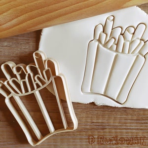 May include: A white plastic cookie cutter shaped like a container of french fries. The cutter is on a wooden surface with a rolling pin in the background.