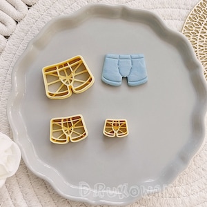 May include: Four cookie cutters in the shape of underwear. Two are yellow and have a grid pattern. One is blue and solid. The smallest cutter is yellow and has a grid pattern.