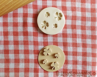 May include: Two white silicone molds with a four-leaf clover design. The molds are on a red and white checkered tablecloth.