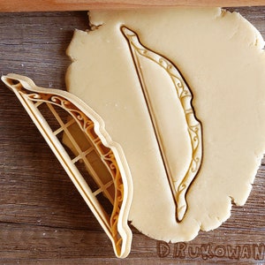 May include: A cookie cutter in the shape of a bow and arrow. The cutter is made of plastic and has a gold color. The cutter is on top of a piece of cookie dough.