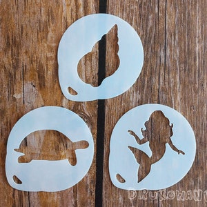 Puede incluir: Tres plantillas de plástico blanco, una con un diseño de concha marina, otra con un diseño de sirena y otra con un diseño de tortuga.
